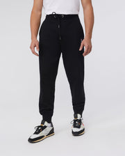 A person stands against a plain background wearing a white t-shirt, classic fit black Psycho Bunny MENS FRENCH TERRY KNIT SWEATPANTS - B6P828ARFT with drawstrings, and black and white sneakers featuring yellow accents.