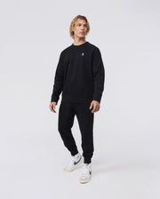 A person in a classic fit black tracksuit and white sneakers stands against a plain background, looking to the side. They wear Psycho Bunny's MENS FRENCH TERRY KNIT SWEATPANTS - B6P828ARFT, which add an effortlessly casual style with their soft French terry fabric.