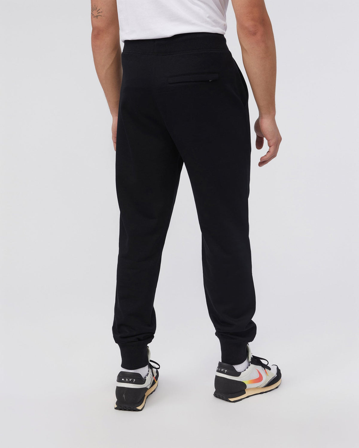A person in Psycho Bunny's MENS FRENCH TERRY KNIT SWEATPANTS (B6P828ARFT), classic fit, with black and white sneakers featuring orange accents, a white T-shirt, and a left-arm tattoo stands facing away. A plain light background highlights the relaxed French terry fabric.