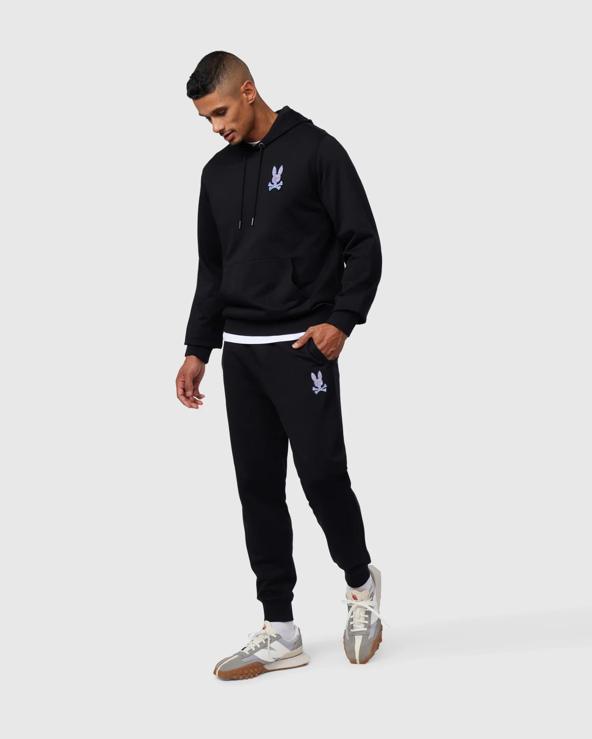 A man is wearing a black hoodie and matching Psycho Bunny MENS SANTA CRUZ SWEATPANT - B6P843A2FT, both made of 100% cotton with a small purple graphic. He is looking down with his left hand in his pocket. He also sports white and gray sneakers with brown soles, exuding comfort and style against a plain gray background.