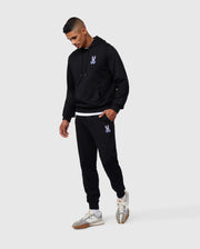 A man is wearing a black hoodie and matching Psycho Bunny MENS SANTA CRUZ SWEATPANT - B6P843A2FT, both made of 100% cotton with a small purple graphic. He is looking down with his left hand in his pocket. He also sports white and gray sneakers with brown soles, exuding comfort and style against a plain gray background.
