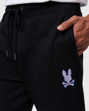 A person's waist and hand wearing black Psycho Bunny MENS SANTA CRUZ SWEATPANT - B6P843A2FT joggers with a drawstring. The joggers, made of 100% cotton for comfort and style, feature a small colorful bunny and crossbones logo on the left hip. The person is wearing a white top.