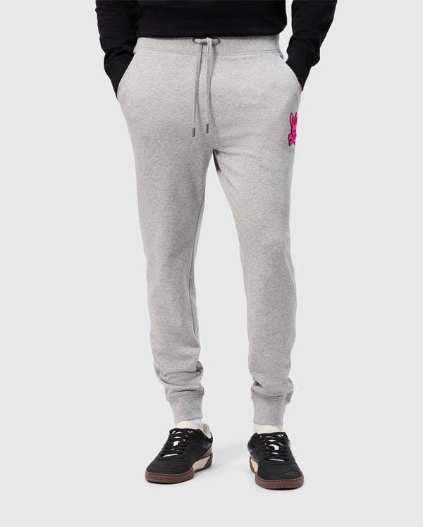 A person wearing Psycho Bunny's light gray SCOTT CHENILLE PATCH SWEATPANT (B6P856D200) with pockets and a pink embroidered logo on the left. They're paired with black sneakers featuring white soles, set against a plain light gray background.