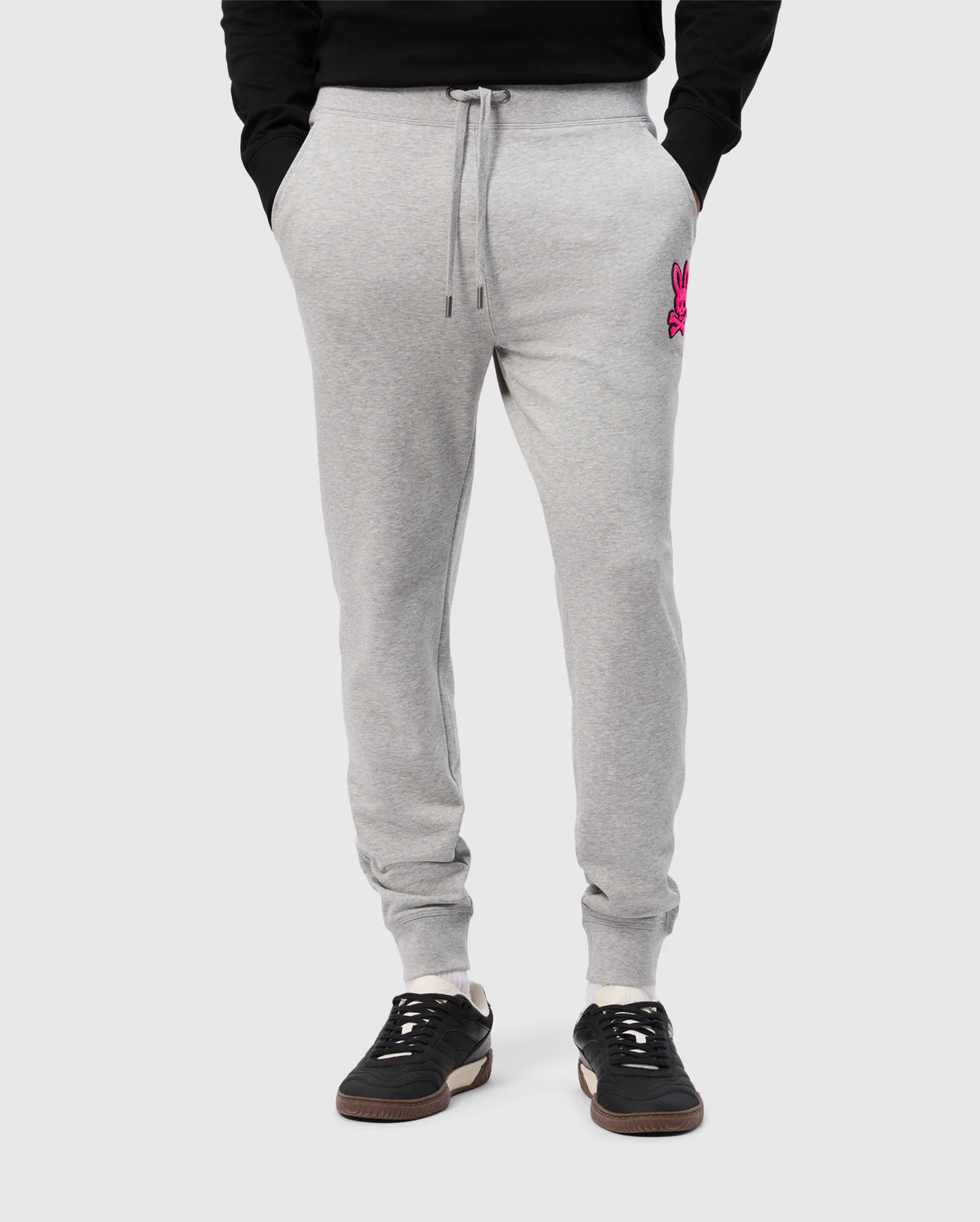 A person wearing Psycho Bunny's light gray SCOTT CHENILLE PATCH SWEATPANT (B6P856D200) with pockets and a pink embroidered logo on the left. They're paired with black sneakers featuring white soles, set against a plain light gray background.