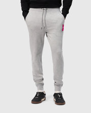 A person wearing Psycho Bunny's light gray SCOTT CHENILLE PATCH SWEATPANT (B6P856D200) with pockets and a pink embroidered logo on the left. They're paired with black sneakers featuring white soles, set against a plain light gray background.
