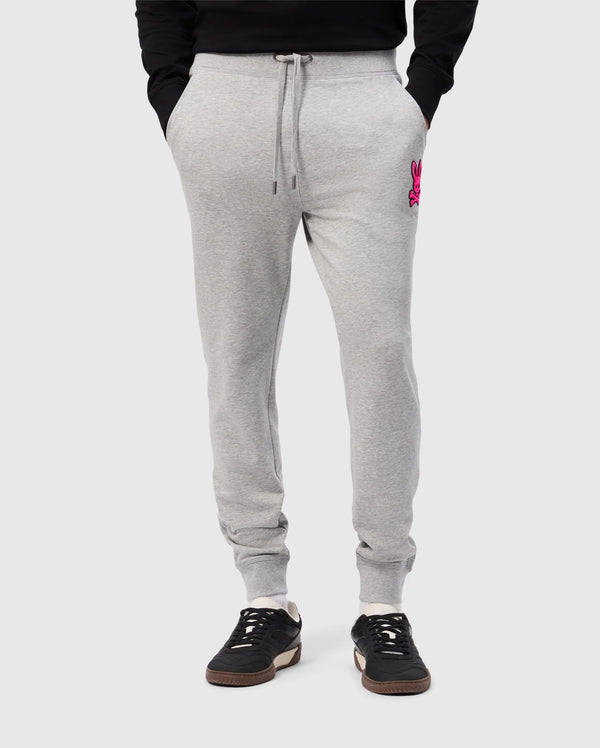 A person wearing Psycho Bunny's light gray SCOTT CHENILLE PATCH SWEATPANT (B6P856D200) with pockets and a pink embroidered logo on the left. They're paired with black sneakers featuring white soles, set against a plain light gray background.