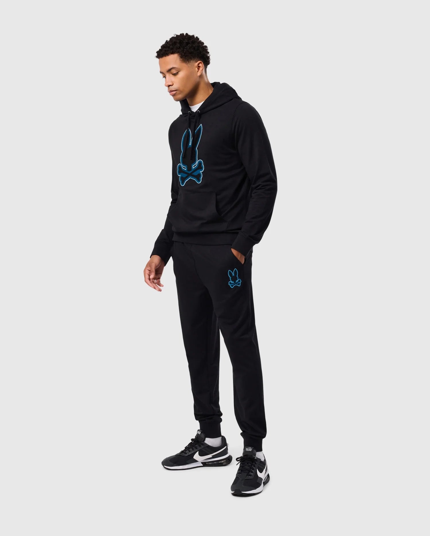 A person dressed in a black hoodie and the MENS WALTER LIGHTWEIGHT SWEATPANT - B6P860D200 from Psycho Bunny, featuring a blue bunny design, stands against a plain gray background. Additionally, the individual wears black sneakers with white soles and slightly looks downwards, perfectly embodying casual wear with a modern silhouette.