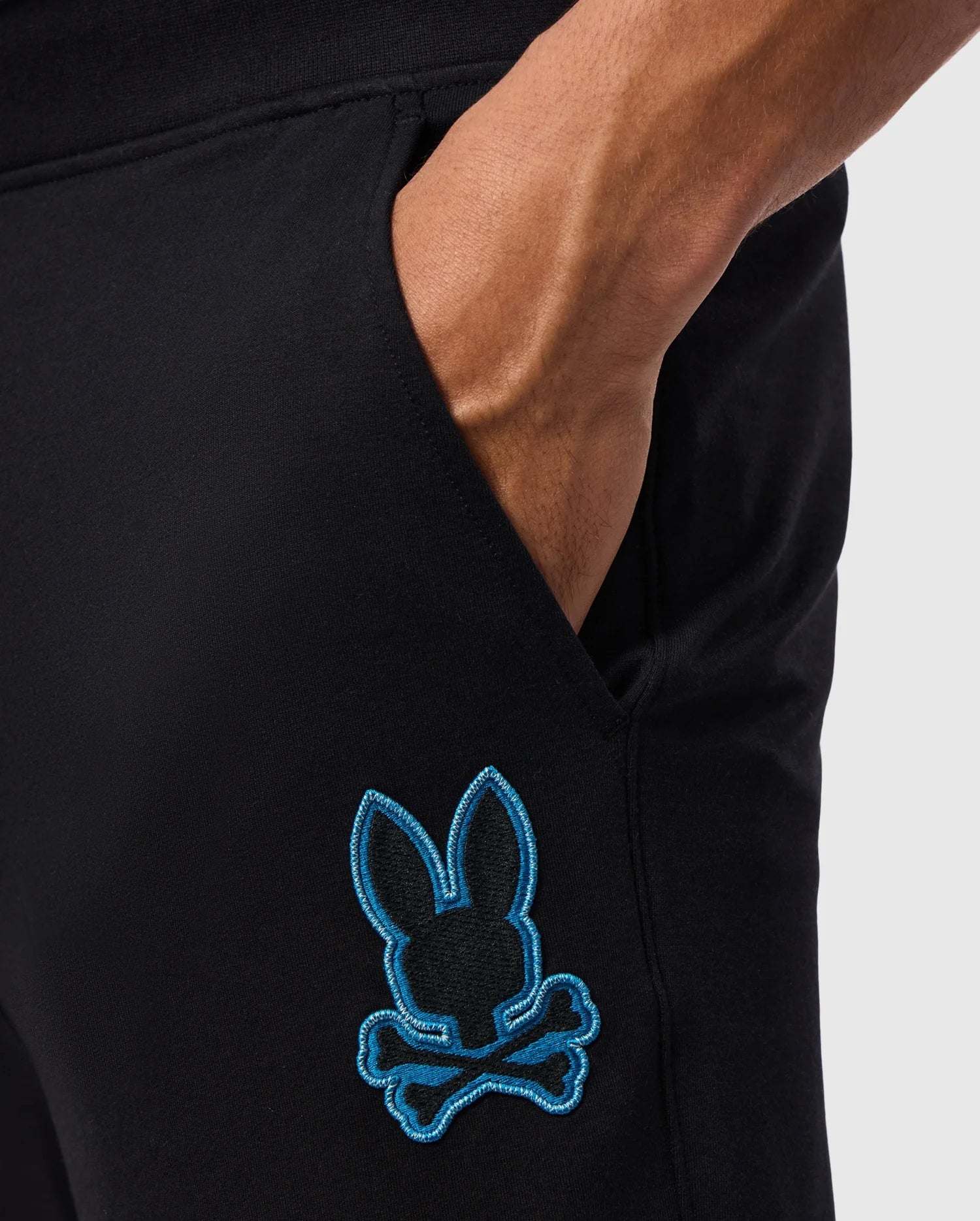 Close-up of a person's hand in the pocket of black MENS WALTER LIGHTWEIGHT SWEATPANT - B6P860D200 by Psycho Bunny. There is an embroidered blue graphic near the pocket featuring a stylized bunny head with crossbones underneath, making these casual wear lightweight sweatpants perfect for effortless style. The background is plain and light-colored.