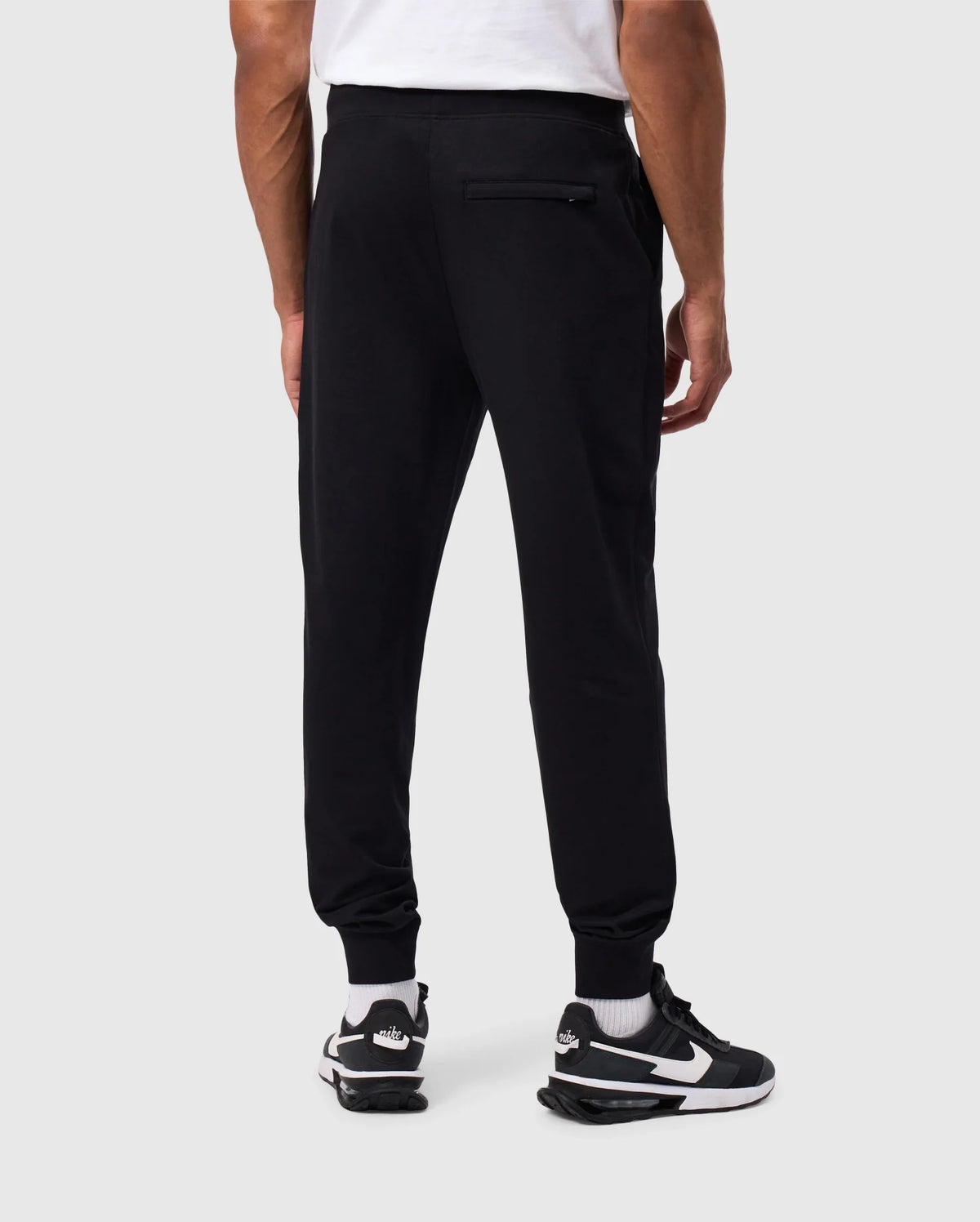 A person is standing with their back to the camera, wearing a white t-shirt, Psycho Bunny MENS WALTER LIGHTWEIGHT SWEATPANT (B6P860D200), and black and white sneakers. The jogger pants, designed for a breathable fit, feature a zippered back pocket. The person is photographed against a plain, light-grey background.