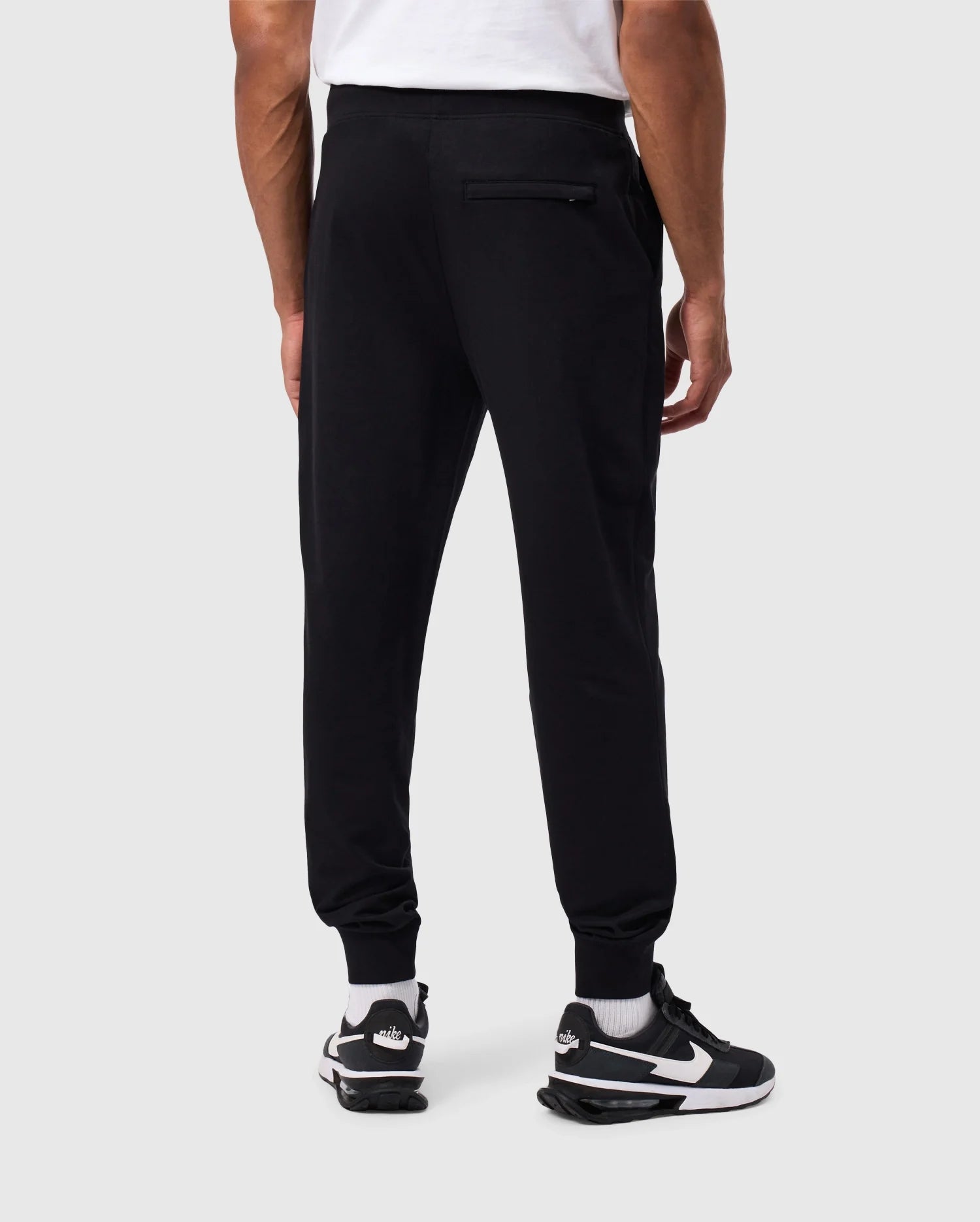 A person is standing with their back to the camera, wearing a white t-shirt, Psycho Bunny MENS WALTER LIGHTWEIGHT SWEATPANT (B6P860D200), and black and white sneakers. The jogger pants, designed for a breathable fit, feature a zippered back pocket. The person is photographed against a plain, light-grey background.