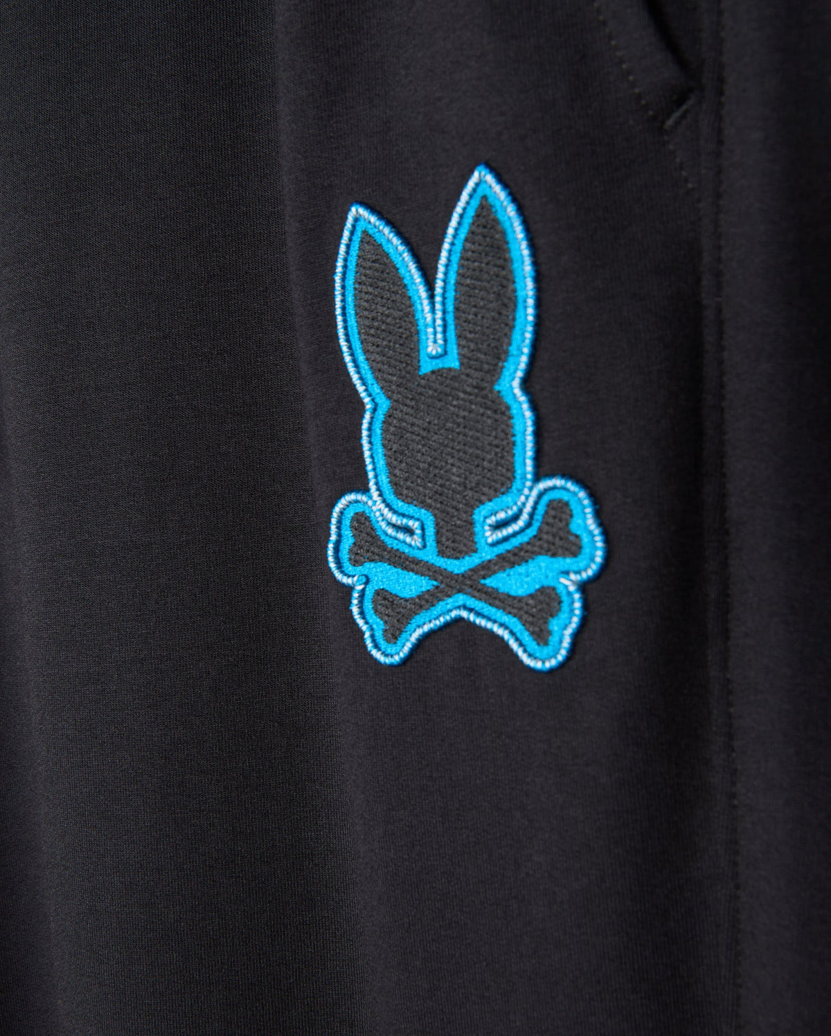The close-up showcases the black fabric of the MENS WALTER LIGHTWEIGHT SWEATPANT - B6P860D200 from Psycho Bunny, featuring a blue embroidered rabbit head above crossed bones, echoing a pirate skull and crossbones motif. The sharp embroidery injects a burst of color and adds a whimsical flair to this casual wear, combining style with breathable comfort.