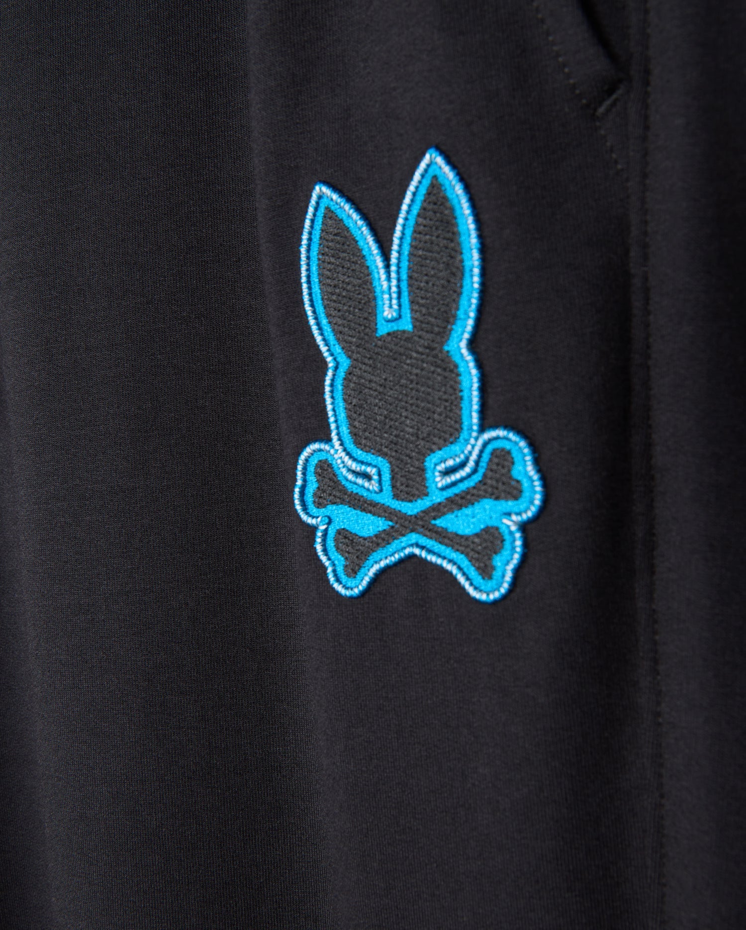 The close-up showcases the black fabric of the MENS WALTER LIGHTWEIGHT SWEATPANT - B6P860D200 from Psycho Bunny, featuring a blue embroidered rabbit head above crossed bones, echoing a pirate skull and crossbones motif. The sharp embroidery injects a burst of color and adds a whimsical flair to this casual wear, combining style with breathable comfort.