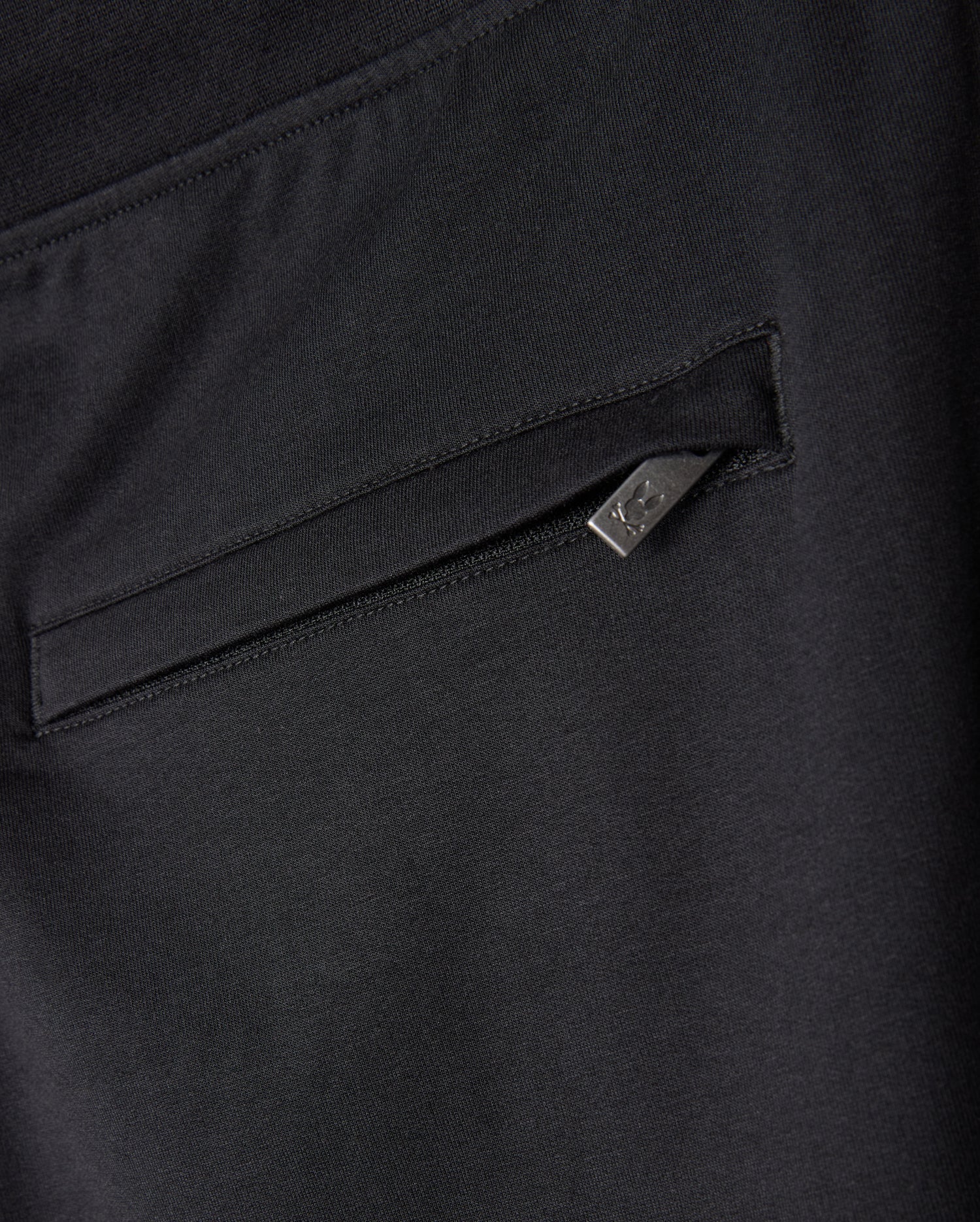 Close-up image of Psycho Bunny's MENS WALTER LIGHTWEIGHT SWEATPANT - B6P860D200 in black, showcasing a zippered side pocket with a small triangular metal tag attached to the zipper. The fabric has a smooth and slightly stretchy texture, ideal for a lightweight sweatpant that blends casual wear with a modern silhouette.
