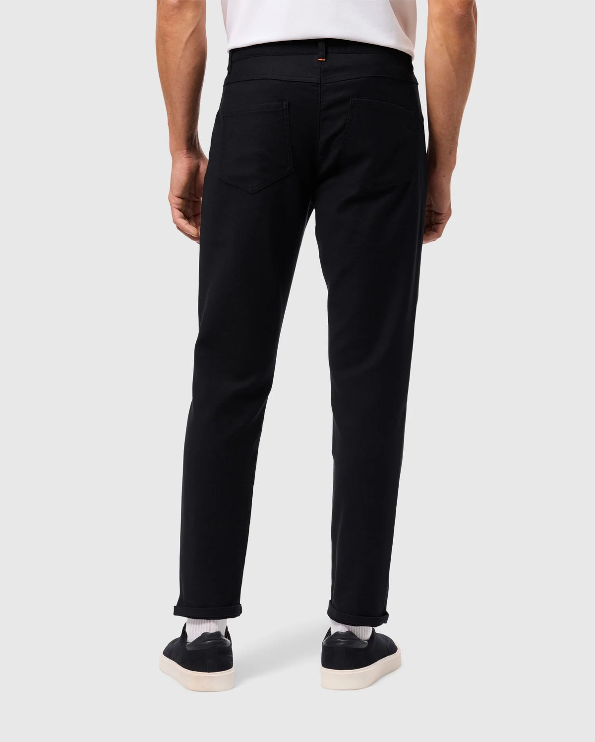 A person wearing a regular fit white shirt, MENS HAYS TWILL PANT by Psycho Bunny in black, and black sneakers with white soles stands with their back facing the camera. The background is a plain light gray.