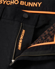 A close-up shot showcases the MENS HAYS TWILL PANT by Psycho Bunny in black, adorned with orange embroidery spelling "Psycho Bunny." Made from a cotton-blend twill with four-way stretch for enhanced comfort, these pants feature a regular fit. A partially open zipper reveals a lining decorated with an orange bunny pattern, complemented by orange trim and detailing.