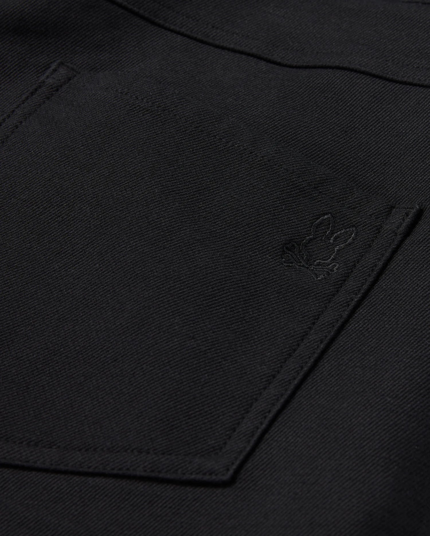 Close-up of Psycho Bunny's MENS HAYS TWILL PANT, showcasing a black fabric featuring a pocket and an embroidered bunny design in the corner. The texture is slightly rough, suggesting a durable cotton-blend twill material.