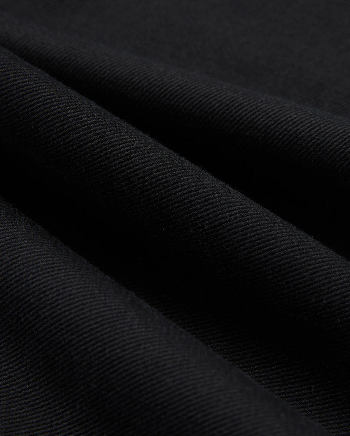 Close-up of Psycho Bunny's MENS HAYS TWILL PANT - B6P894D200, crafted from a black cotton-blend twill fabric with a diagonal ribbed texture. The material boasts four-way stretch, giving it a soft feel and a subtle sheen, with folds producing shadowy areas across the surface.