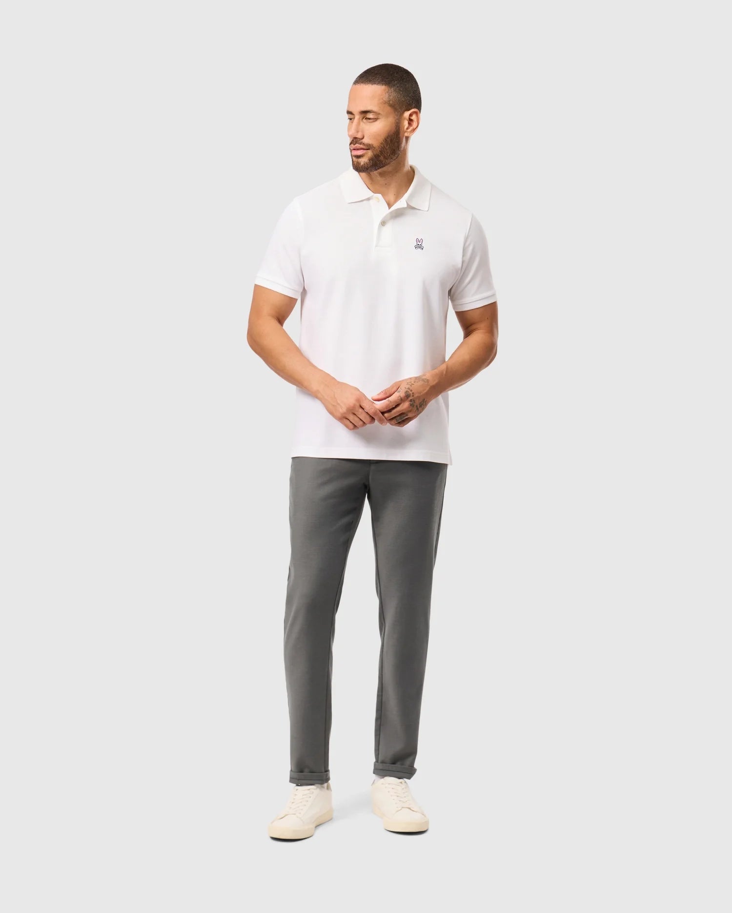 A person wearing a regular fit white polo shirt and Psycho Bunny's gray MENS HAYS TWILL PANT - B6P894D200 stands against a light gray background. They are wearing white shoes, have short hair and a beard, and the polo shirt features a small logo on the chest.