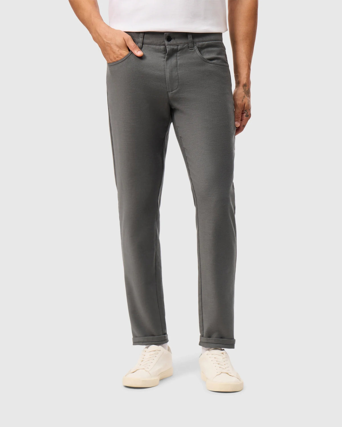 A person wearing a white shirt and the Psycho Bunny MENS HAYS TWILL PANT in gray stands with one hand in their pocket. Paired with white sneakers, these cotton-blend twill jeans embody a casual elegance against the plain background.