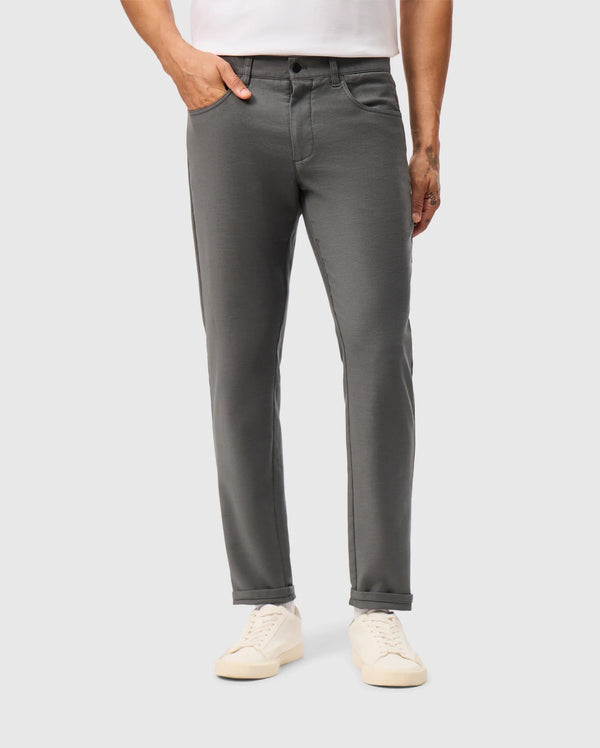 A person wearing a white shirt and the Psycho Bunny MENS HAYS TWILL PANT in gray stands with one hand in their pocket. Paired with white sneakers, these cotton-blend twill jeans embody a casual elegance against the plain background.