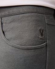 A close-up of the back pocket of the Psycho Bunny MENS HAYS TWILL PANT - B6P894D200 in dark gray cotton-blend twill showcases a small embroidered design featuring a bunny with crossed bones above the pocket. The fabric provides 4-way stretch and has a slightly textured appearance.