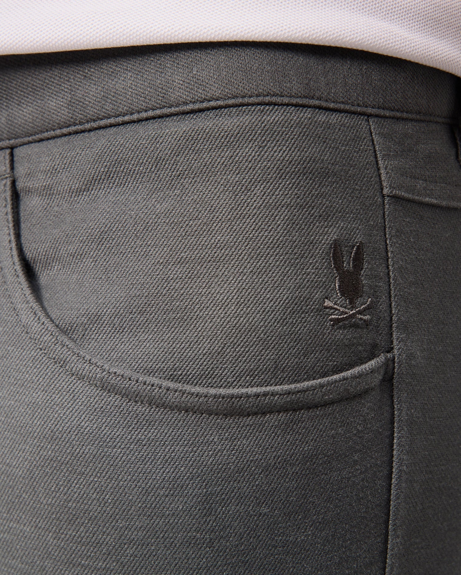 A close-up of the back pocket of the Psycho Bunny MENS HAYS TWILL PANT - B6P894D200 in dark gray cotton-blend twill showcases a small embroidered design featuring a bunny with crossed bones above the pocket. The fabric provides 4-way stretch and has a slightly textured appearance.