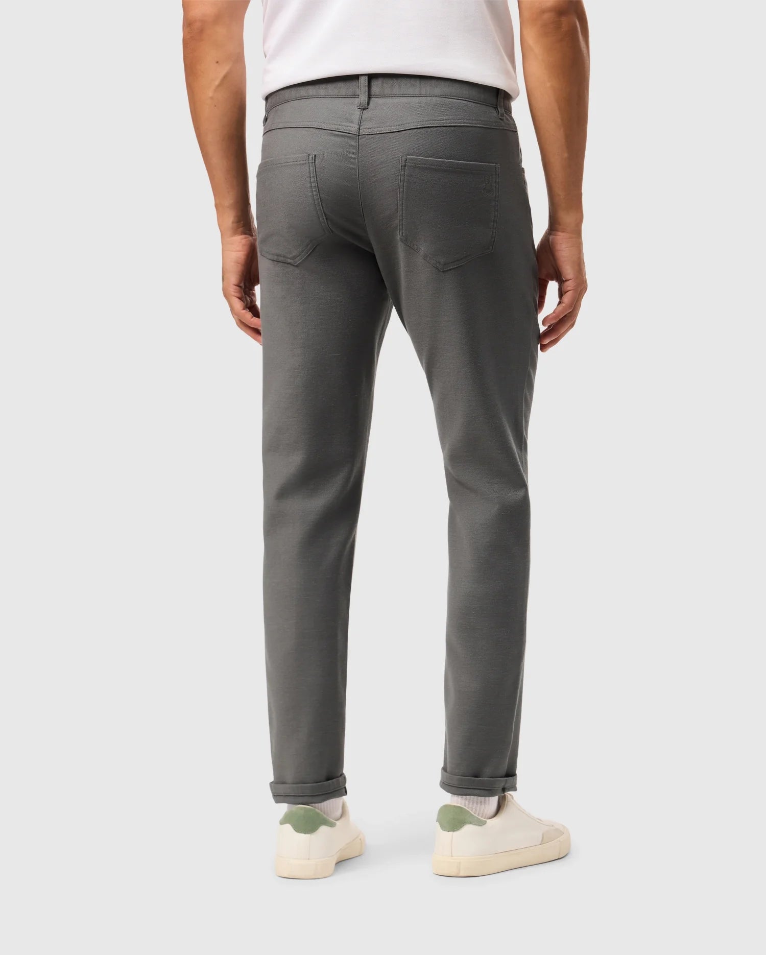A person wearing a white shirt, the Psycho Bunny MENS HAYS TWILL PANT in gray (B6P894D200), and white sneakers with a green accent on the heel stands facing away. The slim-fit pants include back pockets. The background is plain and light-colored.