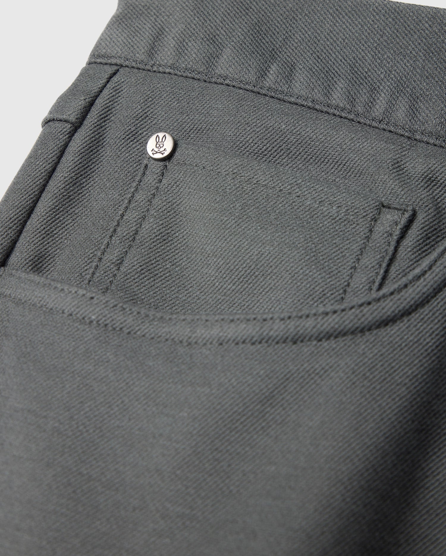 Close-up of the MENS HAYS TWILL PANT - B6P894D200 by Psycho Bunny, showcasing a pocket adorned with a small metallic button engraved with a minimalist face design. Situated on the pocket's upper corner, this dark gray cotton-blend twill garment exhibits 4-way stretch and features a textured, sturdy fabric.
