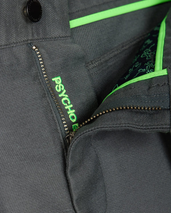 Close-up of the MENS HAYS TWILL PANT by Psycho Bunny, crafted from a gray cotton-blend twill fabric featuring a partially visible zipper. The iconic "PSYCHO" is embroidered in green next to the zipper, with vibrant green accents on the inner fabric flap. The material provides 4-way stretch for enhanced comfort and flexibility.