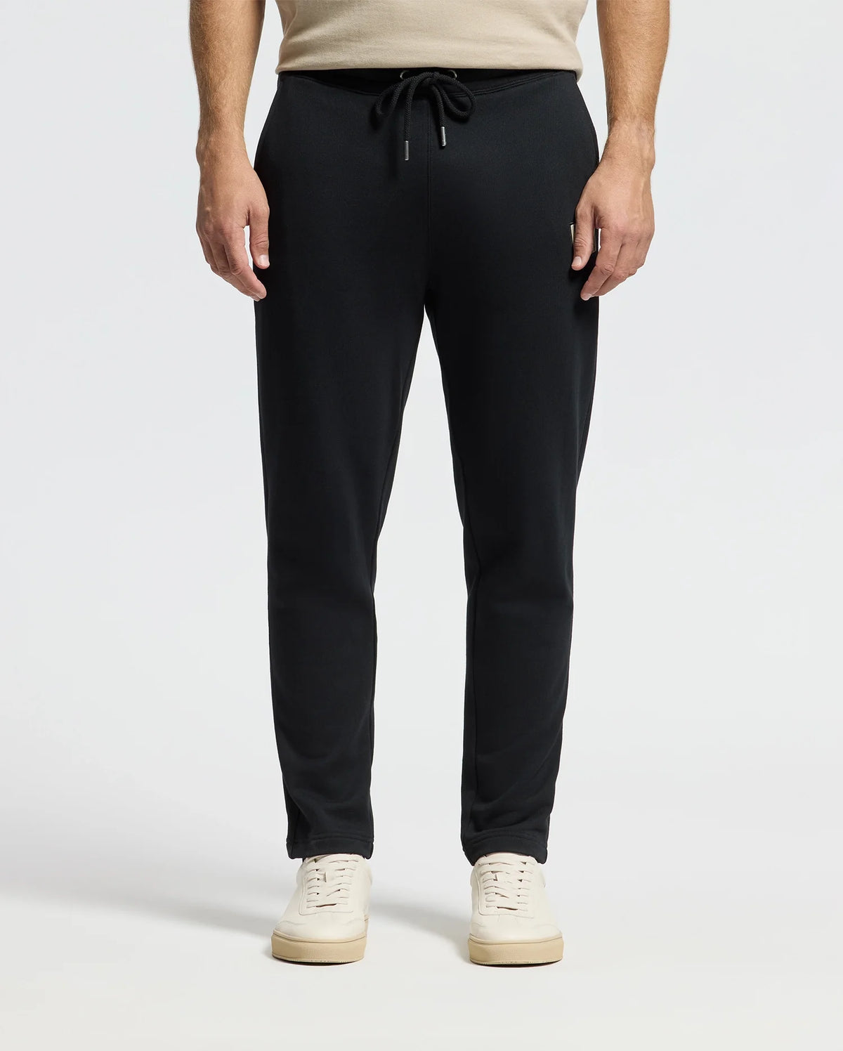 A person wearing black Men's Monroe Twill Patch Fleece Sweatpants by Psycho Bunny and white sneakers stands against a gray background. Their hands rest naturally by their sides, and the drawstring of the 100% cotton pants is tied in a bow at the waist.