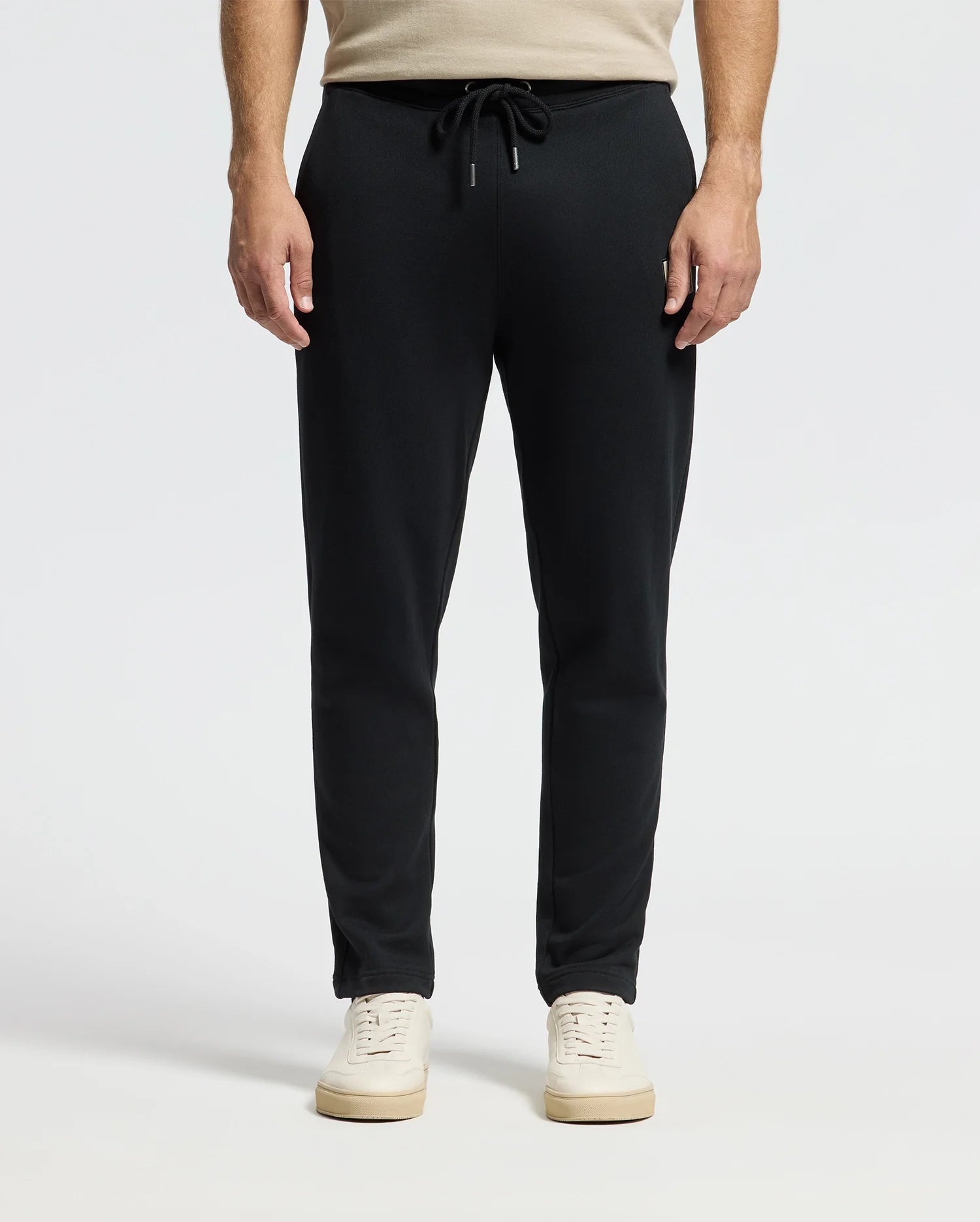 A person wearing black Men's Monroe Twill Patch Fleece Sweatpants by Psycho Bunny and white sneakers stands against a gray background. Their hands rest naturally by their sides, and the drawstring of the 100% cotton pants is tied in a bow at the waist.