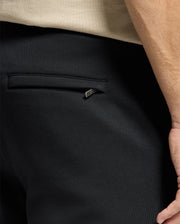 Close-up of a person wearing dark Psycho Bunny MENS MONROE TWILL PATCH FLEECE SWEATPANT - B6P929D200, featuring 100% cotton fabric with a back pocket and a metal zipper pull. The edge of a beige shirt is visible at the top of the image, while the hand rests by the side without any accessories.