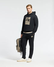 A man stands wearing a black hoodie with a graphic print and Psycho Bunny's MENS MONROE TWILL PATCH FLEECE SWEATPANT, creating a coordinated look. In one hand, he holds a beige and black backpack while sporting white sneakers. The plain, light-colored background emphasizes his stylish 100% cotton outfit.