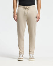 A person wearing the Psycho Bunny MENS MONROE TWILL PATCH FLEECE SWEATPANT - B6P929D200, designed in beige with a soft French terry texture and a subtle embroidered logo on the left pocket, stands against a plain background. The pants are made from 100% cotton, featuring a drawstring waistband for a relaxed fit. They are paired with casual sneakers and a black top.