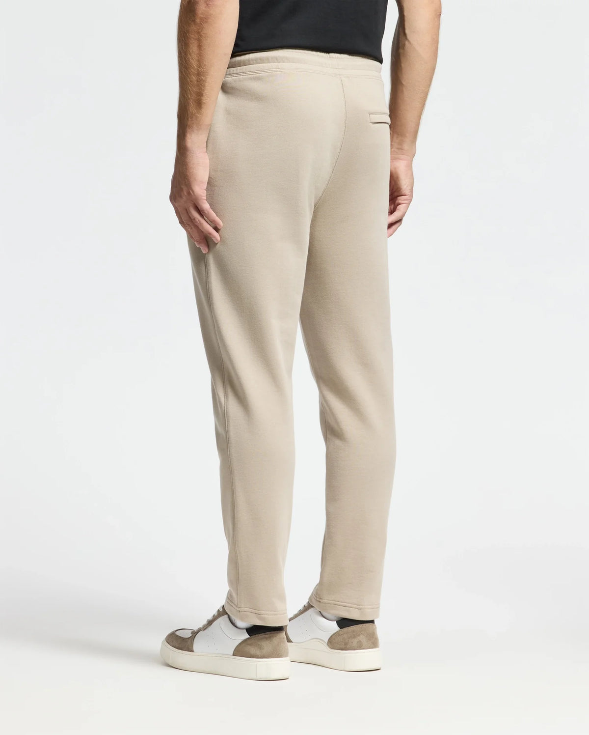 A person dressed in beige Monroe Twill Patch Fleece Sweatpants by Psycho Bunny and white sneakers with brown accents is standing against a plain white background. The image focuses on the lower half of the body from behind, showcasing the casual elegance of 100% cotton comfort.