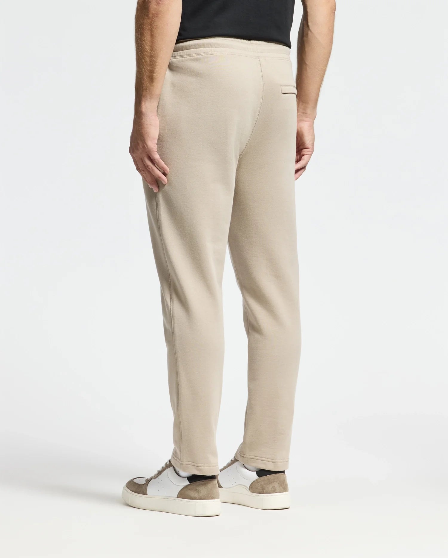 A person dressed in beige Monroe Twill Patch Fleece Sweatpants by Psycho Bunny and white sneakers with brown accents is standing against a plain white background. The image focuses on the lower half of the body from behind, showcasing the casual elegance of 100% cotton comfort.