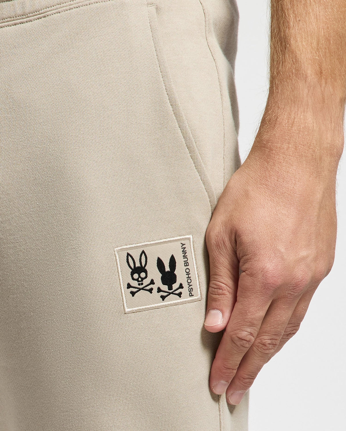 A close-up showcases a person donning the Psycho Bunny MENS MONROE TWILL PATCH FLEECE SWEATPANT - B6P929D200 in beige, crafted from 100% cotton. Their hand rests on the side, and the pants are adorned with a stitched patch of a bunny face and crossbones.