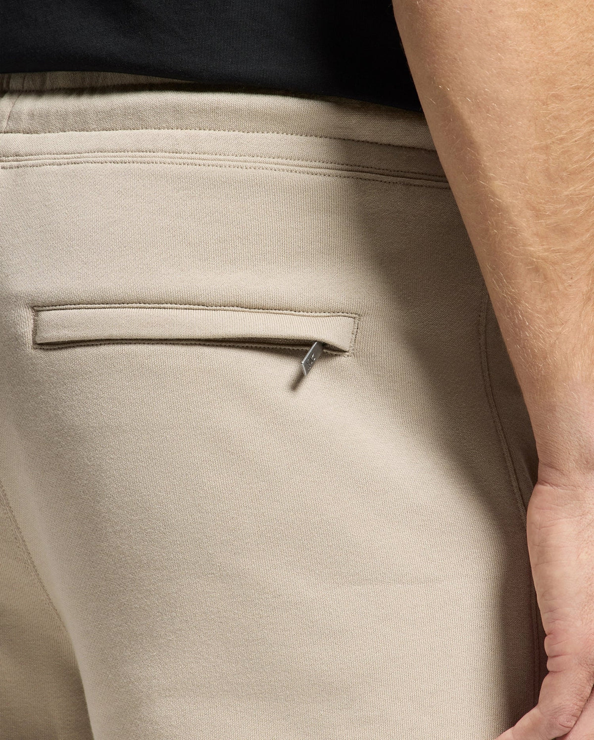 Close-up of a hand resting near the pocket of the beige Mens Monroe Twill Patch Fleece Sweatpant - B6P929D200 by Psycho Bunny. The focus is on the pocket, with a small metal zipper pull slightly exposed. The person is wearing a black top, creating a chic contrast with the comfortable look of 100% cotton fabric.