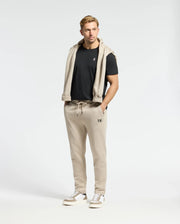 A man stands facing the camera against a plain background, wearing a black French terry t-shirt and beige Psycho Bunny Monroe Twill Patch Fleece Sweatpants with a matching sweatshirt draped over his shoulders. He has short blond hair and white sneakers.