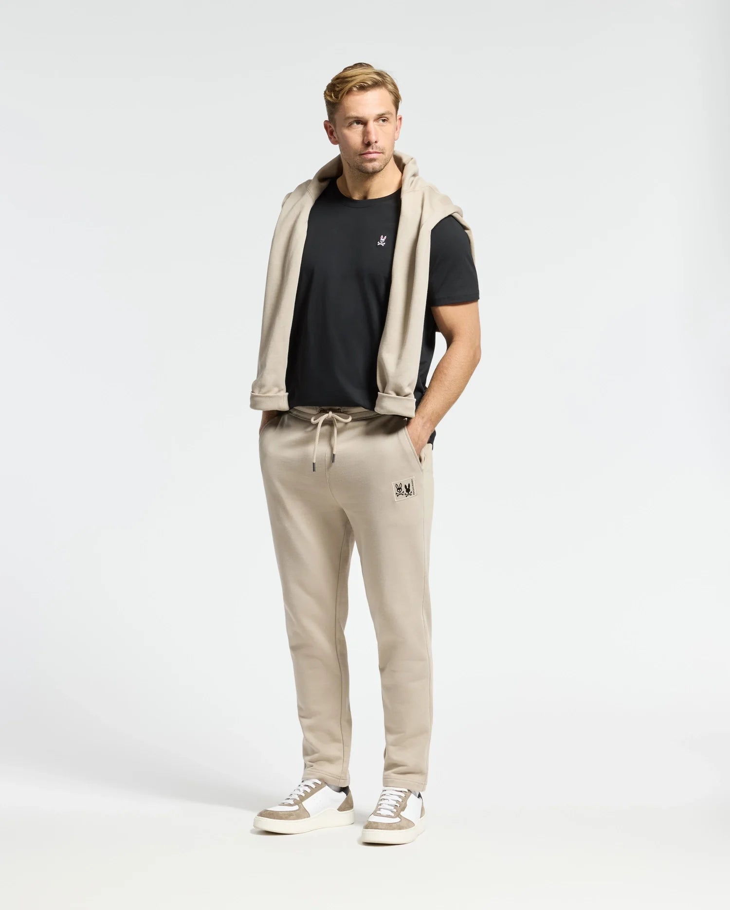 A man stands facing the camera against a plain background, wearing a black French terry t-shirt and beige Psycho Bunny Monroe Twill Patch Fleece Sweatpants with a matching sweatshirt draped over his shoulders. He has short blond hair and white sneakers.