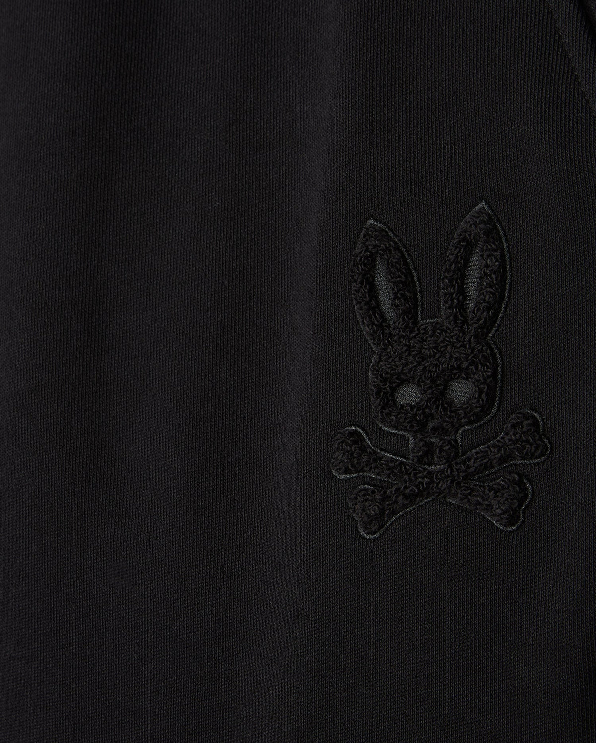 Close-up of the Psycho Bunny MENS PACIFIC CHENILLE JOGGER - B6P972D200 in black, highlighting an embroidered design of a bunny head with crossbones. This casual bottom showcases the stylish high-pile chenille logo patch detail that stands subtly raised from the fabric.