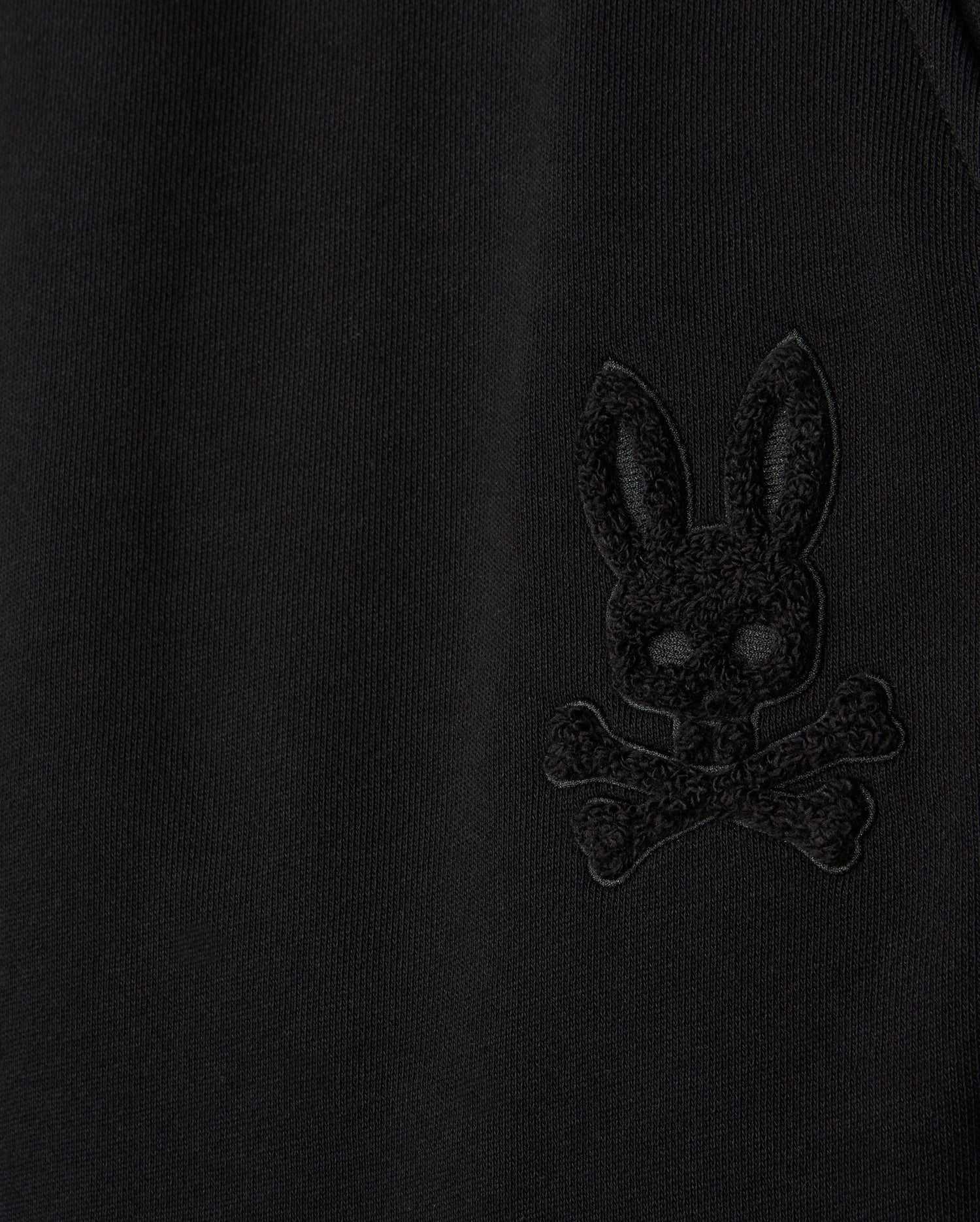 Close-up of the Psycho Bunny MENS PACIFIC CHENILLE JOGGER - B6P972D200 in black, highlighting an embroidered design of a bunny head with crossbones. This casual bottom showcases the stylish high-pile chenille logo patch detail that stands subtly raised from the fabric.