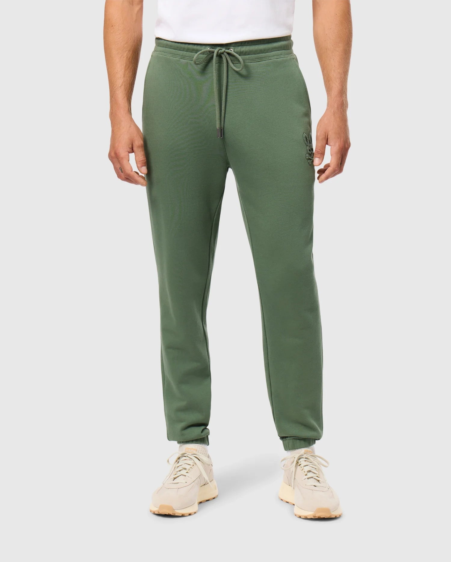 A person wearing Psycho Bunny's MENS PACIFIC CHENILLE JOGGER - B6P972D200, complete with a drawstring, paired with a white shirt and beige sneakers. The image highlights the lower body clad in these casual joggers featuring a high-pile chenille logo patch, set against a plain background.