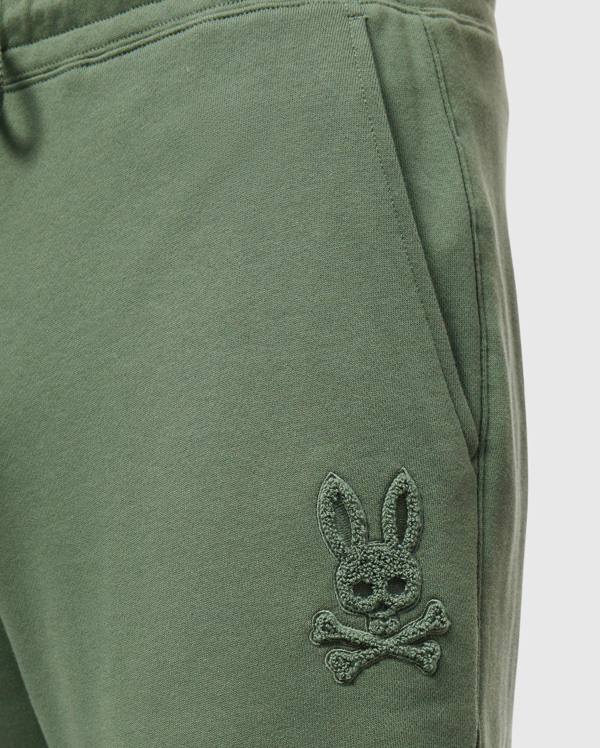 Close-up of green Men's Pacific Chenille Jogger - B6P972D200 from Psycho Bunny, featuring an embroidered logo of a stylized bunny head with crossbones beneath it. The fabric looks soft and comfortable, making it an ideal casual bottom for any relaxed outing.