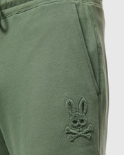 Close-up of green Men's Pacific Chenille Jogger - B6P972D200 from Psycho Bunny, featuring an embroidered logo of a stylized bunny head with crossbones beneath it. The fabric looks soft and comfortable, making it an ideal casual bottom for any relaxed outing.