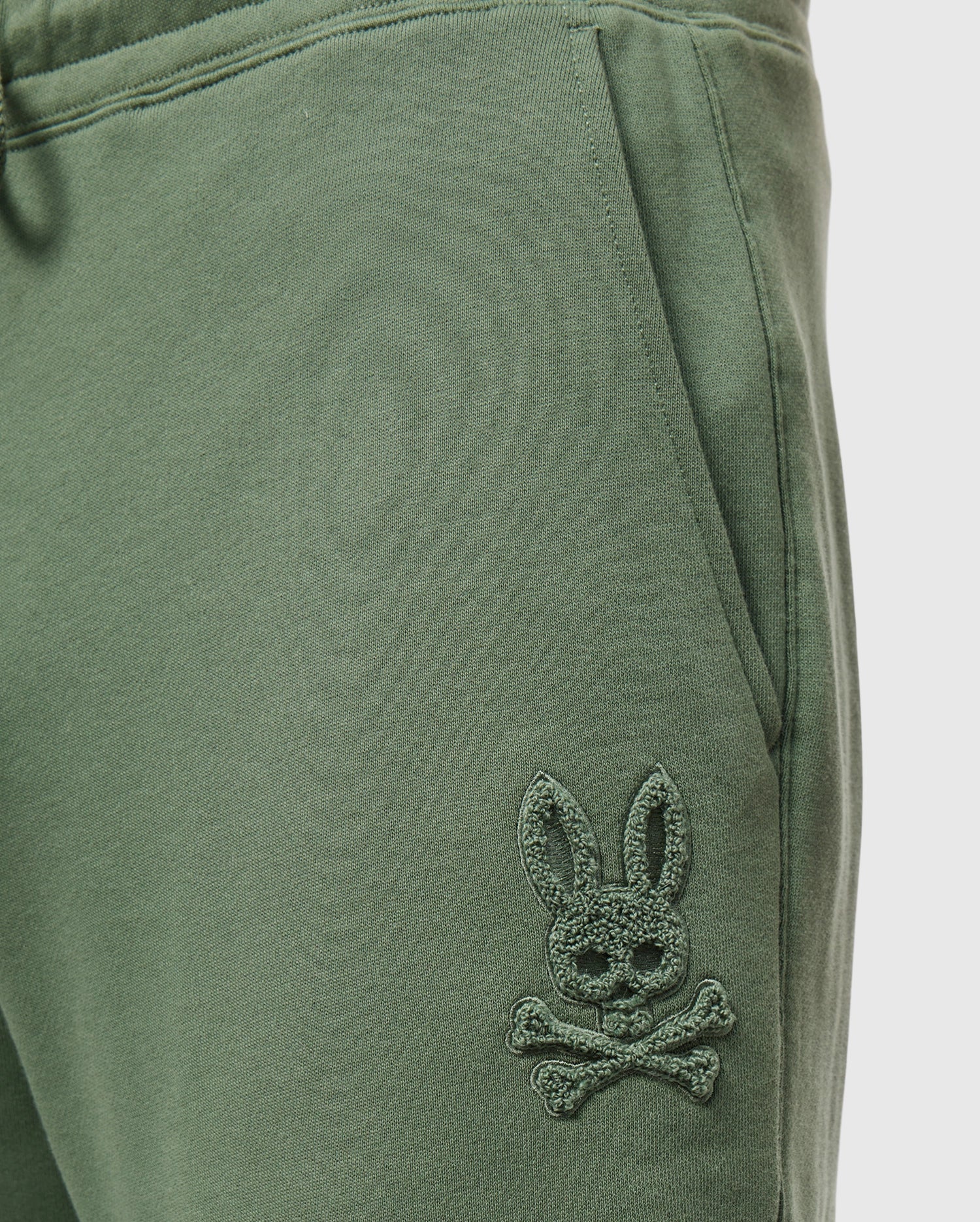 Close-up of green Men's Pacific Chenille Jogger - B6P972D200 from Psycho Bunny, featuring an embroidered logo of a stylized bunny head with crossbones beneath it. The fabric looks soft and comfortable, making it an ideal casual bottom for any relaxed outing.