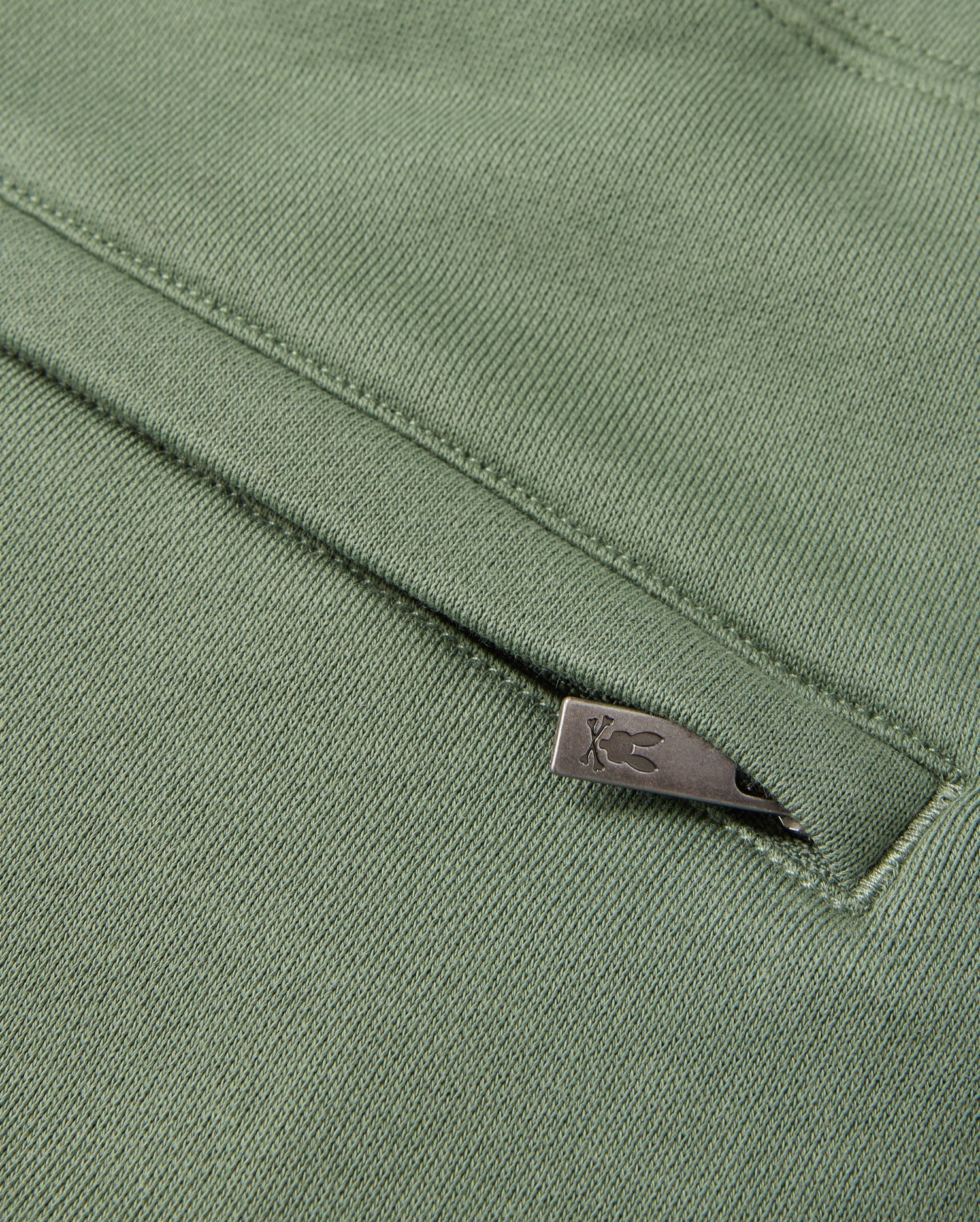 Close-up of a textured, green fabric featuring a sleek metallic zipper partially visible. The smooth fabric showcases intricate stitching details, reminiscent of the high-pile chenille logo patch found on the MENS PACIFIC CHENILLE JOGGER by Psycho Bunny.
