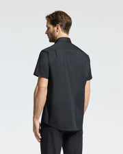 A man with short brown hair stands facing away on a plain white background, wearing the sleek Psycho Bunny MENS GABRIEL SHORT SLEEVE SHIRT - B6Q351F200 in woven stretch fabric, paired with black pants.