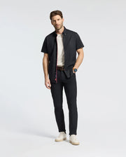 A person stands casually against a light gray background, wearing a Psycho Bunny MENS GABRIEL SHORT SLEEVE SHIRT over a white T-shirt, black pants, white sneakers, and a watch. One hand is in their pocket while the other hangs by their side.