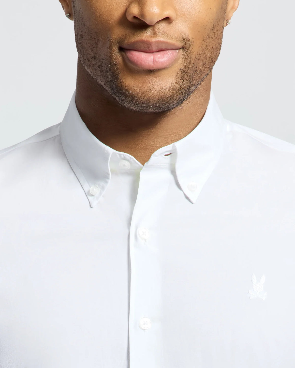 A person in a Psycho Bunny white Gabriel short sleeve shirt, made from luxurious Pima cotton with an embroidered Bunny logo on the chest. The image shows the upper torso and face with a neutral expression, featuring a neatly buttoned classic collar for a polished look.